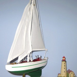 May include: A white model sailboat with green and red accents on a white stand. The sailboat has a white sail and is displayed next to a miniature lighthouse.