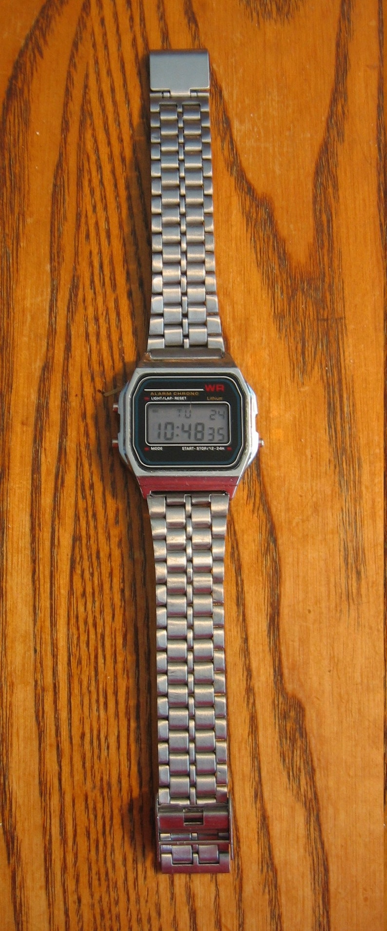 Puede incluir: Un reloj de pulsera digital plateado con una esfera rectangular y una correa de metal. El reloj muestra la hora 10:48:35. El reloj tiene el texto "WR" y "ALARM CHRONO" en la esfera.