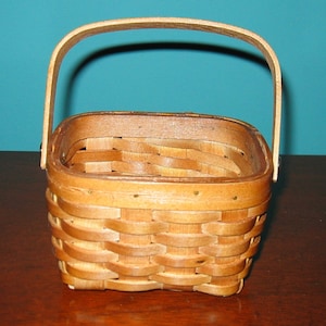 May include: A small, square, woven basket with a wooden handle. The basket is made of natural wood and has a light brown color.