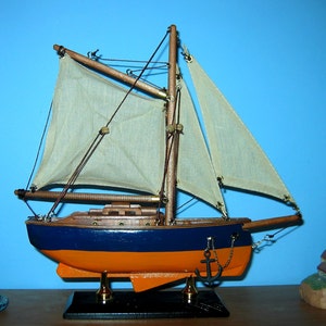 May include: A wooden model sailboat with white sails, a blue and orange hull, and a black base. The sailboat is on a shelf with two miniature lighthouses.