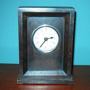 May include: A dark wood framed clock with a white face and black hands. The clock face has Roman numerals and the text "Westminster Time" in a cursive font.
