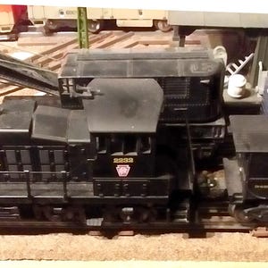 Può includere: Una locomotiva ferroviaria modello nera Pennsylvania con un accessorio gru e un vagone di coda abbinato. La locomotiva è contrassegnata con il testo PENNSYLVANIA in lettere gialle e il numero 8833. Un vagone blu e bianco è visibile sullo sfondo.