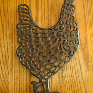 May include: A black cast iron chicken silhouette with a decorative, lacy design. The chicken is standing on a wooden surface.