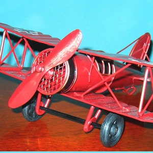 May include: A red metal model airplane with a propeller. The airplane has a biplane design with two sets of wings. The model is detailed with a black and gold engine and a white star on the tail.