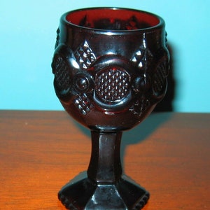 Puede incluir: Un cáliz de vidrio rojo oscuro con un diseño estampado. El cáliz tiene una copa ancha y un tallo alto y delgado.
