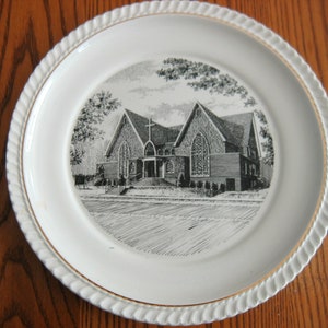 Könnte beinhalten: Ein weißer Keramikteller mit goldfarbenem Rand zeigt eine Schwarzweißillustration eines Kirchengebäudes. Die Kirche hat ein Kreuz auf dem Dach und ist von Bäumen umgeben.
