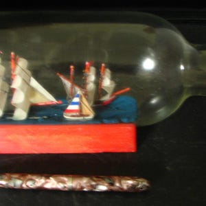 May include: A clear glass bottle containing a miniature model ship with white sails and red masts, resting on a red wooden base with a cork stopper. Below the bottle lies a metallic, patterned pen with a silver tip, providing a scale reference.