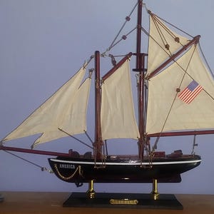 May include: A model ship with white sails and a black hull, with the word "America" on the side. The ship is on a wooden base with a gold nameplate that reads "Schooner." The ship is displayed next to two miniature lighthouses.