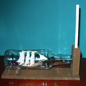 May include: A clear glass bottle with a wooden base and a small ship inside. The bottle is held upright by a wooden stand with a white marker attached to it. The marker has the text "Harbor Bay Craft Village" printed on it.