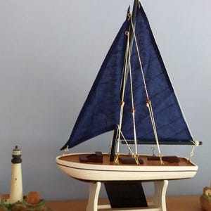 May include: A model sailboat with a white hull, a wooden deck, and dark blue fabric sails, supported by a white stand. Flanking the sailboat are two miniature lighthouse figurines, each featuring a tower and small buildings on a rocky base.
