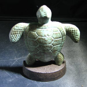 May include: A bronze-colored turtle figurine with outstretched flippers and a detailed shell pattern. The sculpture is mounted on a dark, circular base. The turtle's color has a greenish hue, suggesting an aged or weathered appearance.