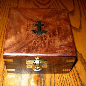 Puede incluir: Una caja de madera con un cierre de lat&oacute;n y un dise&ntilde;o de ancla de metal en la parte superior. La caja tiene una veta de madera marr&oacute;n oscura.