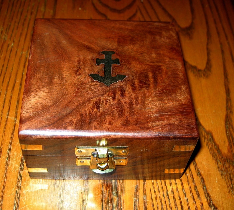 Puede incluir: Una caja de madera con un cierre de lat&oacute;n y un dise&ntilde;o de ancla de metal en la parte superior. La caja tiene una veta de madera marr&oacute;n oscura.