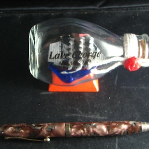 May include: A clear glass bottle with the words "Lake George NEW YORK" printed on it, resting on an orange stand. The bottle is sealed with white string and a red wax seal. A brown and copper-coloured pen lies below.