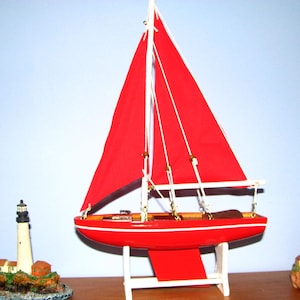 May include: A model sailboat with a bright red hull and matching red fabric sails, mounted on a white wooden stand. Flanking the sailboat are two miniature lighthouse figurines, each featuring a tall tower and small buildings on a rocky base.