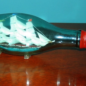 May include: A miniature ship model inside a clear glass bottle with a red stopper. The bottle is on a wooden surface.