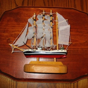 May include: A wooden model ship with white sails, a black and red hull, and a wooden base. The ship is displayed on a wooden plaque.