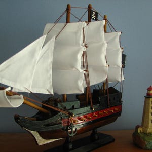 May include: A detailed wooden model of a pirate ship with white sails and a black Jolly Roger flag. The ship has a black hull with red accents and is displayed next to a miniature lighthouse.