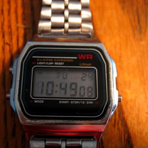 Puede incluir: Un reloj digital plateado con una esfera negra que muestra la hora 10:49:08. El reloj tiene una esfera rectangular con el texto "WR" y "Lithium". La correa del reloj est&aacute; hecha de eslabones plateados.