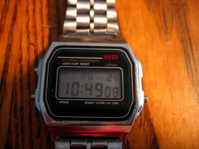 Puede incluir: Un reloj digital plateado con una esfera negra que muestra la hora 10:49:08. El reloj tiene una esfera rectangular con el texto "WR" y "Lithium". La correa del reloj est&aacute; hecha de eslabones plateados.
