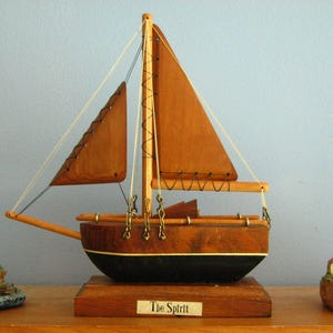 May include: A wooden sailboat model with brown sails and a black hull, labeled "The Spirit," is flanked by two decorative lighthouse figurines. The lighthouses are white and beige with red tops, set on miniature landscapes.