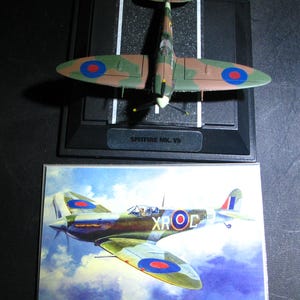May include: A model of a Spitfire Mk. Vb airplane, featuring a camouflage pattern of green and brown. The model sits on a black base with white runway markings. A picture of the same plane is below the model.