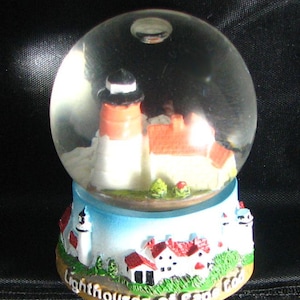 Puede incluir: Un globo de nieve con una escena de faro. El globo tiene un faro blanco y naranja, edificios con tejados rojos y arbustos verdes. La base es azul y tiene las palabras "Lighthouses of Cape Cod".