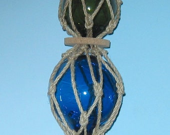 May include: Three glass fishing floats, each encased in a net of natural twine. The top float is green, the middle float is blue, and the bottom float is amber.