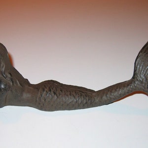 May include: A dark grey cast iron mermaid figurine, lying down with her tail extended. The mermaid has long hair and is detailed with textured scales and fins. This decorative item is suitable for home decor.