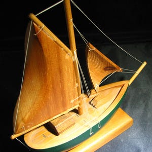 May include: A wooden model of a sailing boat with tan sails and a green hull. The model sits on a rectangular wooden base. The sails are secured with white string, and the boat features a detailed wooden deck.