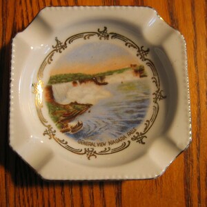 Puede incluir: Cenicero de cerámica blanca con borde dorado y una imagen de las Cataratas del Niágara. El texto "General View Niagara Falls" está impreso en dorado en el cenicero.