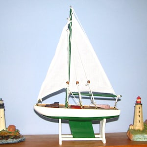 May include: A white-sailed sailboat model with green and white accents, displayed on a white stand. Two lighthouse figurines flank the sailboat, one on each side. The lighthouses have tan and red details.
