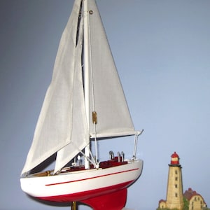 May include: A model sailboat with white sails and a red and white hull. The sailboat is displayed on a white base with a gold-colored support. A small lighthouse figurine is to the right of the sailboat.