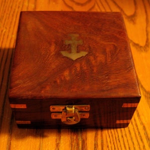 Puede incluir: Una caja de madera con un dise&ntilde;o de ancla de lat&oacute;n en la tapa. La caja tiene un pestillo y bisagras de lat&oacute;n.
