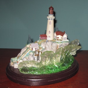 Può includere: Una statuetta di faro in miniatura con una torre bianca e un tetto rosso, circondata da onde verdi e una piccola casa. Il faro è posto su una base marrone.