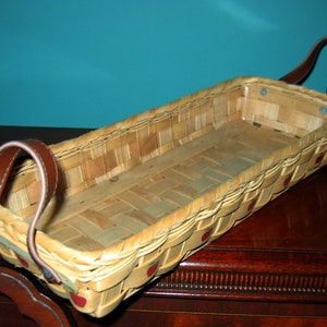 May include: A rectangular woven basket with brown leather handles. The basket has a light brown color and is decorated with red painted apples.