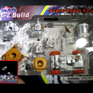 Op de afbeelding: Een doos met plastic modelbouwsets voor het bouwen van een maanwagen, astronauten en andere ruimteverkenningsapparatuur. De doos heeft een rood en gouden ontwerp met de woorden "INATR E-Z Build" en "Scale Model Kits".