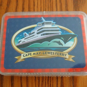 Könnte beinhalten: Eine blaue und rote Karte mit einem weißen Fährschiff darauf. Das Schiff befindet sich in einem goldenen Oval mit den Worten "Cape May-Lewes Ferry" auf einem goldenen Banner.