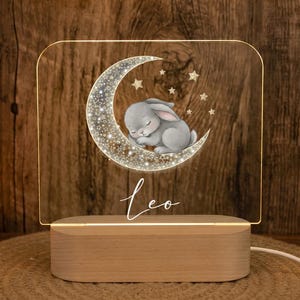 Könnte beinhalten: Ein rechteckiges Acryl-Nachtlicht mit einem aufgedruckten Motiv eines grauen Hasen, der auf einer Mondsichel zwischen Sternen schläft. Der Name Leo ist in weißer Schreibschrift unter dem Mond abgebildet. Das Acrylpanel steht auf einem hellen Holzsockel.