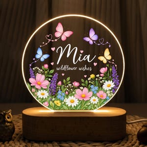 May include: A circular acrylic night light with a warm glow, featuring a printed design of colorful wildflowers, butterflies, and small hearts. The text reads Mia and wildflower wishes in white script. It sits on a light-colored wooden base.