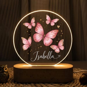 May include: A circular acrylic night light with a warm glow, featuring a printed design of five pink butterflies and gold glitter accents. The name Isabella is written in white cursive script at the bottom, set on a rectangular wooden base.