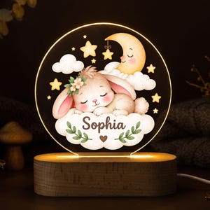 May include: A circular acrylic night light featuring a sleeping bunny on a cloud, a crescent moon, and stars. The name Sophia is printed in dark script on the cloud. The design is illuminated and mounted on a rectangular wooden base.