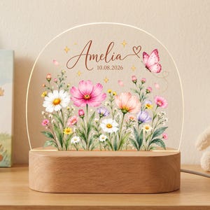 May include: A semi-circular acrylic night light featuring a printed floral garden with pink, white, and purple wildflowers, a pink butterfly, and the name Amelia with the date 10.08.2026. The acrylic panel sits on a smooth, oval wooden base.