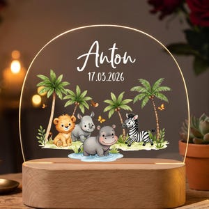 Könnte beinhalten: Ein halbkreisförmiges Acryl-Nachtlicht mit einer aufgedruckten Dschungelszene, die einen Löwen, ein Nashorn, ein Flusspferd und ein Zebra unter Palmen zeigt. Der Text "Anton" und "17.03.2026" steht darüber. Die Acrylplatte ruht auf einem hellen Holzsockel.