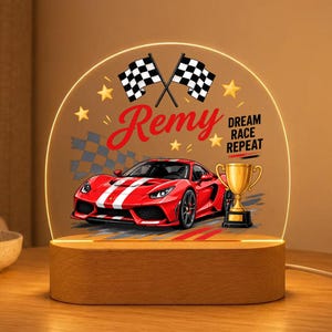 May include: A semi-circular acrylic night light with a wooden base featuring a red sports car, a gold trophy, two checkered flags, and the text "Remy" and "DREAM RACE REPEAT" printed in red and black against a background of yellow stars.
