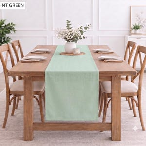 Peut inclure: Un chemin de table rectangulaire de couleur vert menthe en tissu, disposé au centre d'une table à manger en bois. Le chemin de table présente une couleur unie et une texture lisse, s'étendant sur toute la longueur de la surface de la table.