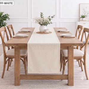 Puede incluir: Un camino de mesa rectangular de color blanco roto hecho de tela, colocado a lo largo de una mesa de comedor de madera. El camino presenta un borde limpio y dobladillado con una textura tejida suave y uniforme.