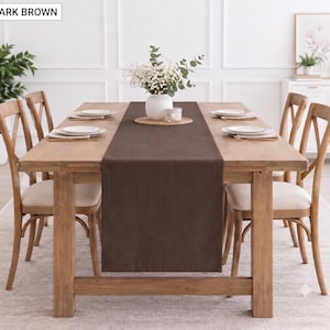 May include: A dark brown rectangular table runner centred on a light wood dining table. The runner features a solid dark brown colour and is positioned beneath a white vase filled with white flowers and a small glass candle holder.