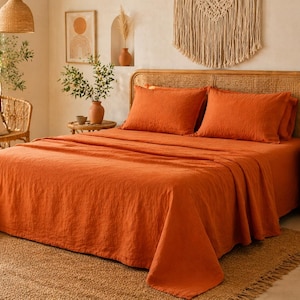 Könnte beinhalten: Ein orangefarbenes Leinen-Bettwäsche-Set, bestehend aus einem Spannbetttuch, einem Bettlaken und zwei Kissenbezügen, wird auf einem Bett mit einem gewebten Kopfteil präsentiert. Der Raum hat eine neutrale Farbpalette mit natürlichen Texturen und Dekorationen.
