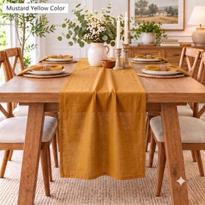 May include: A mustard yellow linen table runner draped over a wooden dining table. The table is set with white plates, mustard yellow napkins, woven placemats, and a white ceramic vase filled with green eucalyptus branches and white flowers.